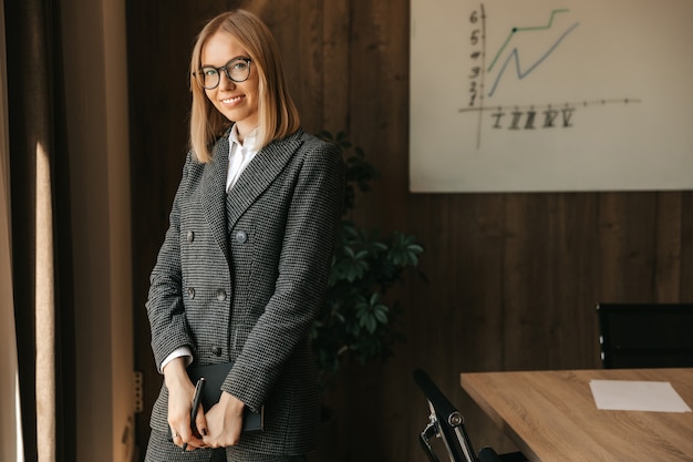 Une belle jeune femme de race blanche à lunettes dans un costume et une chemise se tient près d'un bureau, tenant un cahier dans ses mains et souriant avec un sourire à pleines dents dans le bureau.