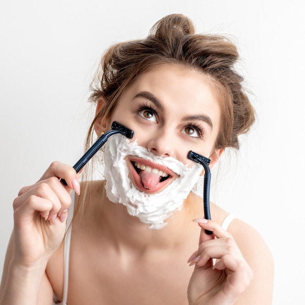 Belle jeune femme de race blanche se raser le visage par deux rasoirs avec la langue sur le mur blanc.