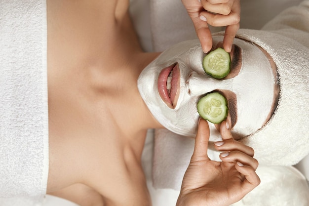 Belle jeune femme reçoit un masque facial à l'argile au spa allongé avec des concombres sur les yeux