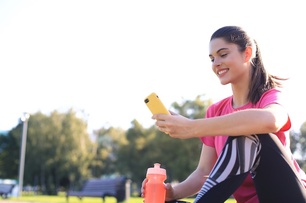 Belle jeune femme regardant loin alors qu'elle était assise sur un tapis d'exercice au parc, se relaxant après l'entraînement et utilisant un smartphone.