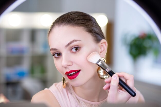 Belle jeune femme regardant son visage tout en mettant des cosmétiques décoratifs