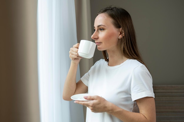 Une belle jeune femme regarde par la fenêtre et boit du café le matin.