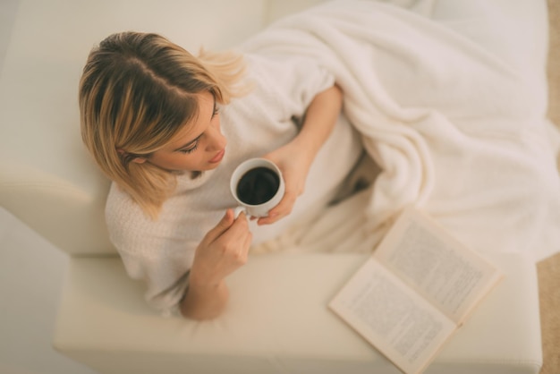 Belle jeune femme relaxante avec une tasse de café et lisant un livre.
