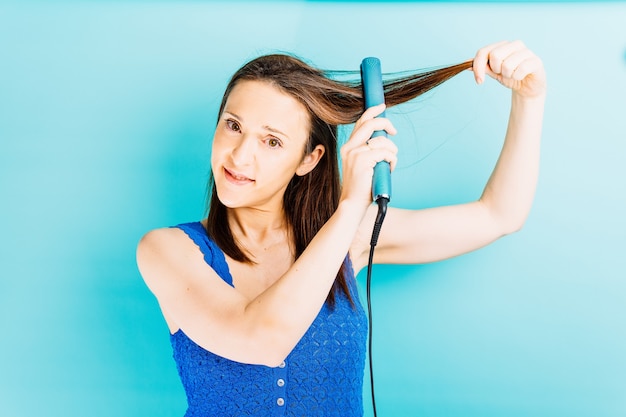 Belle jeune femme repassant ses cheveux avec un fer à lisser sur fond bleu