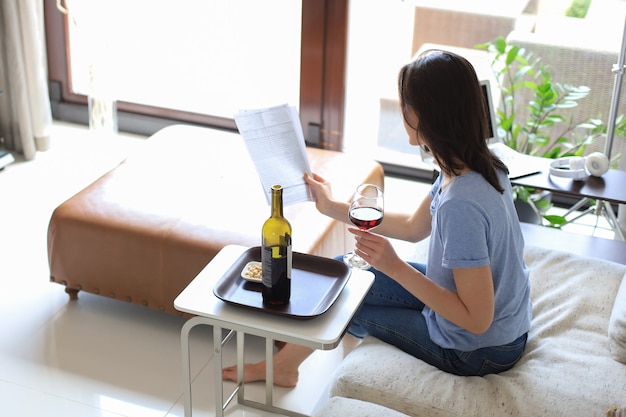 Belle jeune femme réussie assise sur un canapé dans le salon, buvant du vin rouge, lisant des documents.