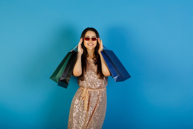 Belle jeune femme en robe beige avec des lunettes rouges tenant de nombreux sacs après des achats réussis.