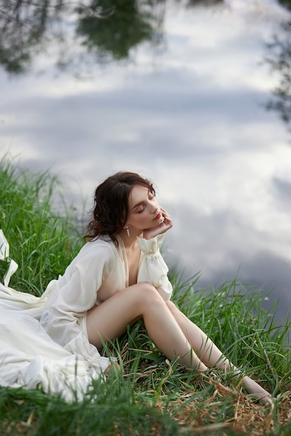 Belle jeune femme en robe blanche reposant sur la rive du lac étang rivière. Maquillage et coiffure professionnels aux cheveux bouclés