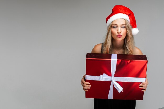 Belle jeune femme en robe et chapeau de Noël détient cadeau et sourires, photo isolée sur mur gris