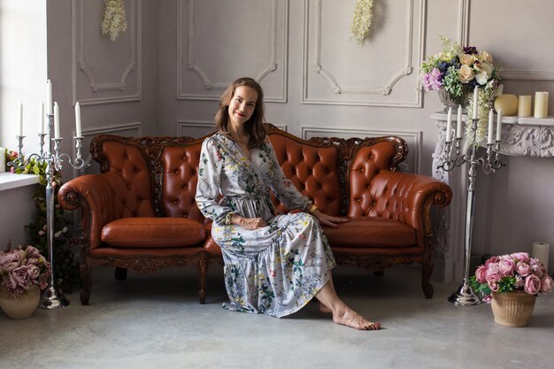 Belle jeune femme en robe à fleurs est assise sur un canapé en cuir marron