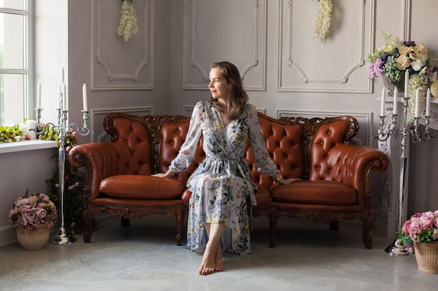 Belle jeune femme en robe à fleurs est assise sur un canapé en cuir marron