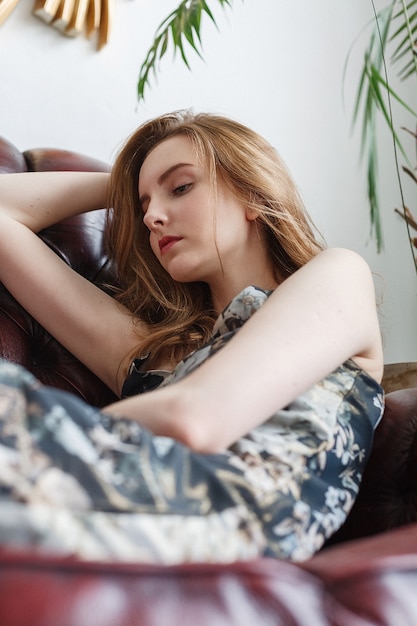 Belle jeune femme en robe hétéroclite, portrait à l'intérieur du modèle réfléchi mignon. assis dans un fauteuil dans une chambre confortable. jolie dame naturelle posant à la maison