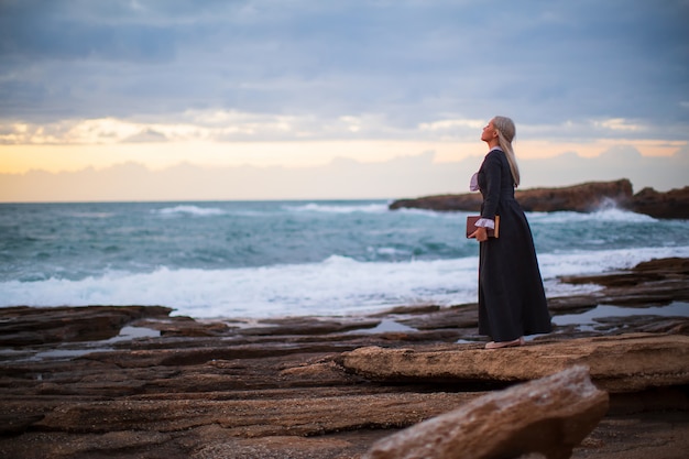 La belle jeune femme romantique regarde le coucher du soleil avec un livre à la main à l'extérieur