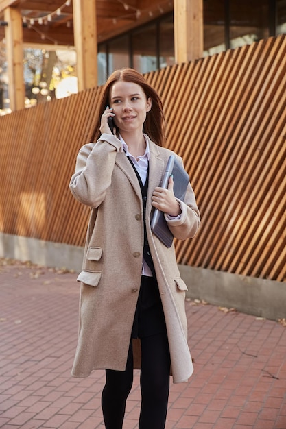 Belle jeune femme rousse caucasienne en manteau beige aux cheveux longs tenant un ordinateur portable à l'extérieur