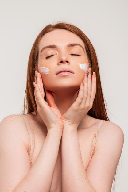 Belle jeune femme rousse dans un soutien-gorge beige fait des soins de la peau et applique de la crème sur son visage