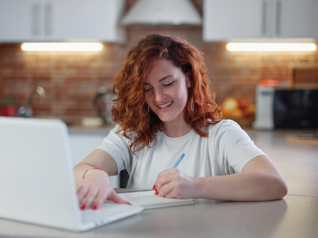 Une belle jeune femme rousse est assise dans la cuisine à table avec son ordinateur portable