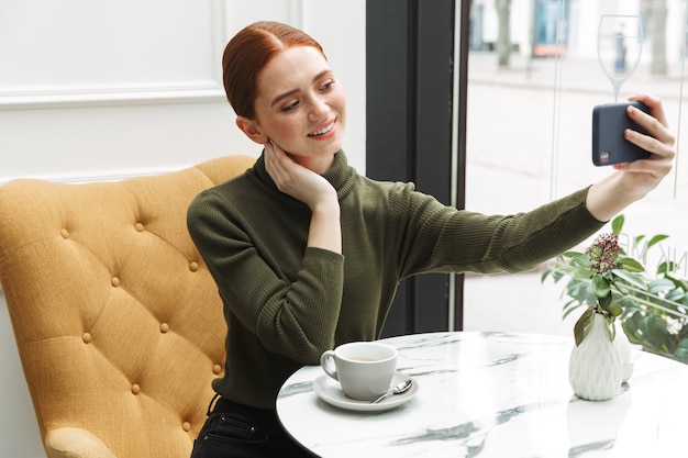 Belle jeune femme rousse se reposant à la table du café à l'intérieur, buvant du café, prenant un selfie