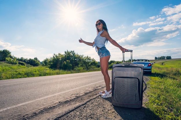 Belle jeune femme sur route rurale avec valise faisant de l'auto-stop sur fond de paysage extérieur journée ensoleillée. La femme a tendu la main avec un doigt vers le haut pour attraper la voiture.