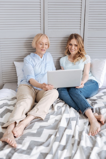 Belle jeune femme et sa mère âgée assise sur le lit et étant entièrement concentrée sur le visionnage de vidéos sur un ordinateur portable