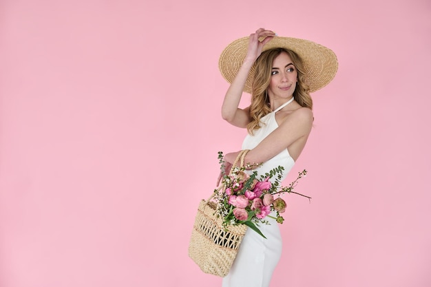 Belle jeune femme avec un sac à main en osier de fleurs dans ses mains et un chapeau de paille sur fond rose