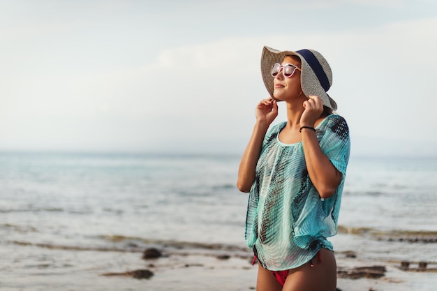 Une belle jeune femme se détend sur la plage par une journée ensoleillée. Elle pose et regarde loin avec le sourire sur son visage.