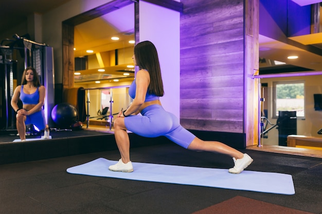 Belle jeune femme se réchauffe et s'étire dans la salle de gym, sur un tapis près du miroir