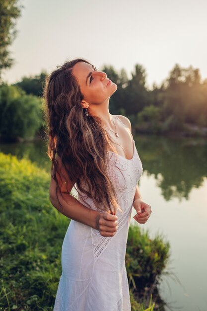 Belle jeune femme se sentant libre et heureuse de se promener au bord de la rivière l'été au coucher du soleil en admirant le paysage