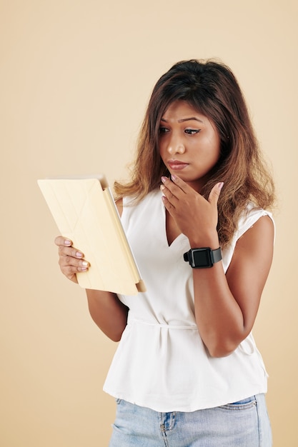 Belle jeune femme sérieuse couvrant la bouche avec la main lors de la lecture de nouvelles choquantes sur une tablette numérique