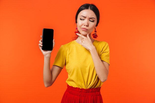 belle jeune femme sérieuse posant isolée sur un mur orange montrant l'affichage du téléphone portable.