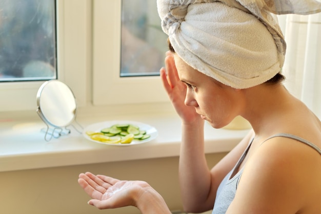 Belle jeune femme avec une serviette de bain sur la tête assise près de la fenêtre de la maison, regardant dans un miroir de maquillage, appliquant de la crème pour le visage. Sur une assiette de rebord de fenêtre avec des tranches de concombre et de citron, des cosmétiques naturels