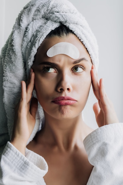 Belle jeune femme en serviette blanche et robe avec des pansements oculaires sur son front posant avec grimace