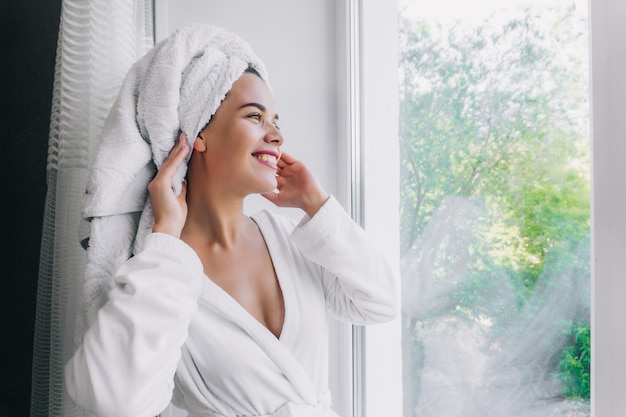 Belle jeune femme en serviette blanche et robe se verrouillant par la fenêtre à la maison