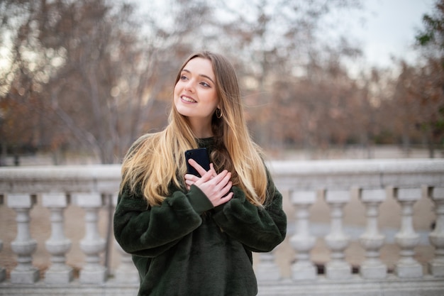 Belle jeune femme souriant avec amour et regardant loin tout en se tenant dans un parc et tenant un smartphone près de la poitrine