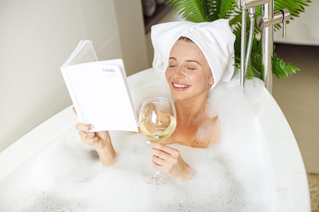 Belle jeune femme souriante allongée dans un bain avec un livre de lecture en mousse et buvant du vin