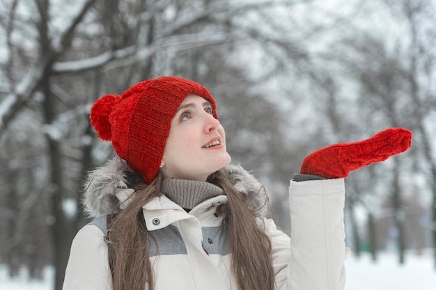 Belle jeune femme souriante au bonnet rouge tend sa paume dans le gant dans les bois. Promenez-vous dans le parc enneigé.
