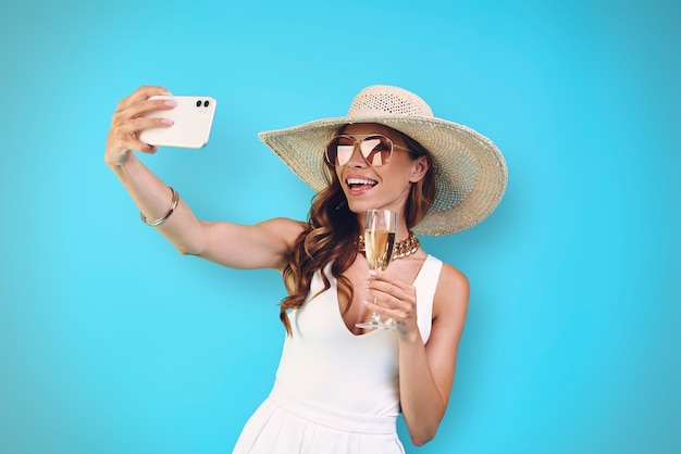 Belle jeune femme souriante au chapeau élégant tenant une flûte à champagne et faisant du selfie