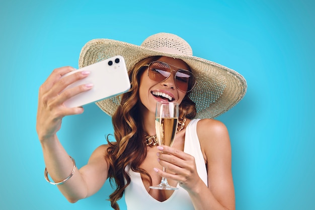 Belle jeune femme souriante au chapeau élégant tenant une flûte à champagne et faisant du selfie