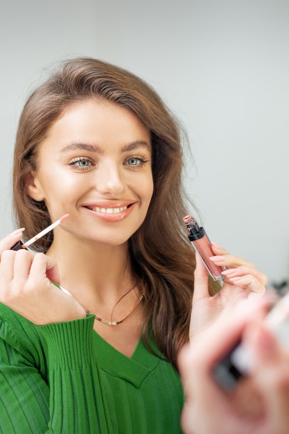 Belle jeune femme souriante caucasienne appliquant le brillant sur les lèvres en regardant dans le miroir