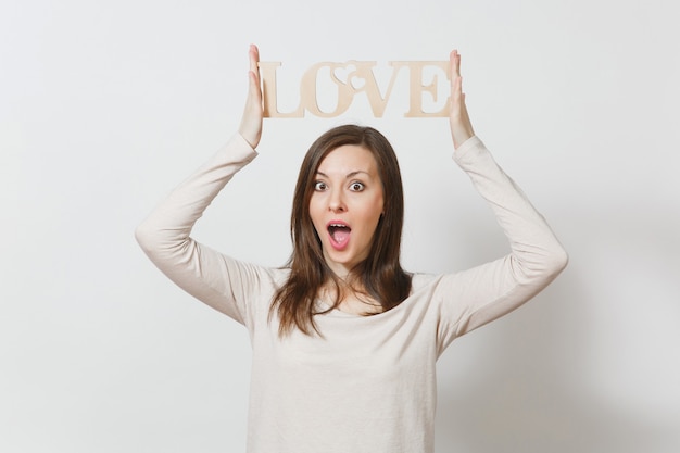 Belle jeune femme souriante dans des vêtements légers et décontractés tenant au-dessus de sa tête amour de mot en bois sur fond blanc. Copiez l'espace pour la publicité. Concept de la Saint-Valentin ou de la Journée internationale de la femme