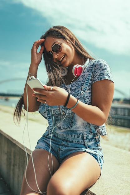 Une belle jeune femme souriante envoie des SMS sur son smartphone ou utilise les médias sociaux et profite de la rive de la rivière de la ville par une belle journée d'été.