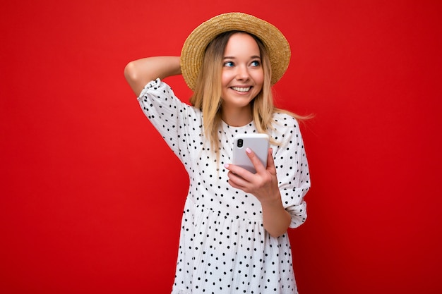 Belle jeune femme souriante et heureuse portant des vêtements décontractés, isolée sur fond, surfant sur Internet par téléphone en regardant sur le côté