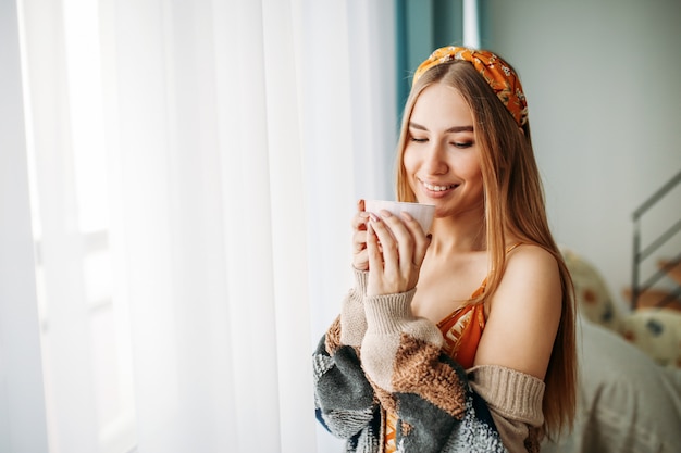 Belle jeune femme souriante juste cheveux longs fille portant un cardigan tricoté confortable avec une tasse de thé près de la fenêtre à la maison