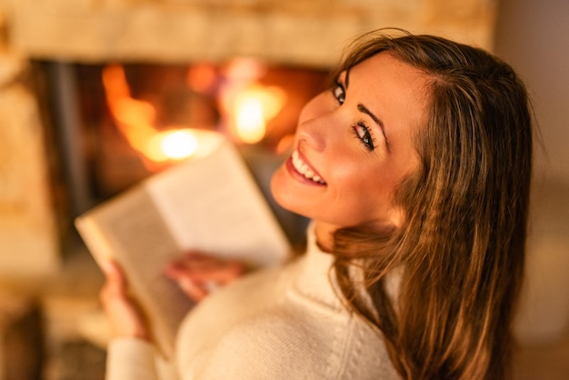 Belle jeune femme souriante lisant un livre et profitant de la cheminée.