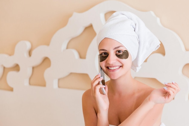 Belle jeune femme souriante avec des patchs sous les yeux et téléphone portable parlant en peignoir assis dans son lit