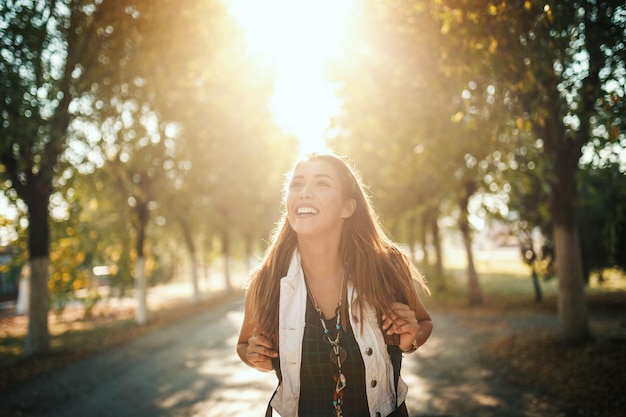 Une belle jeune femme souriante avec des sacs à dos sur le dos marche le long de l'avenue ensoleillée d'automne et regarde au loin.