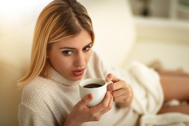 Belle jeune femme souriante se détend sur son canapé dans le salon en buvant du café et en lisant un livre.