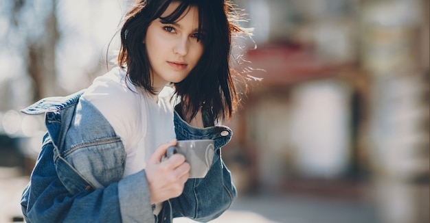 Belle jeune femme souriante tout en tenant une tasse de café en touchant ses cheveux à l'extérieur dans un café.