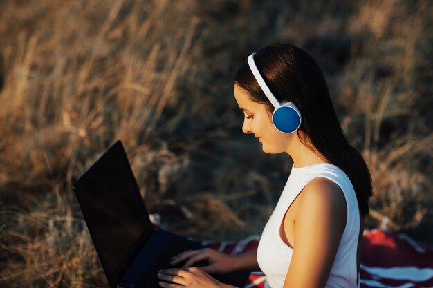 Belle jeune femme souriante travaillant sur ordinateur portable et porter des écouteurs alors qu'il était assis sur l'herbe dans le parc.