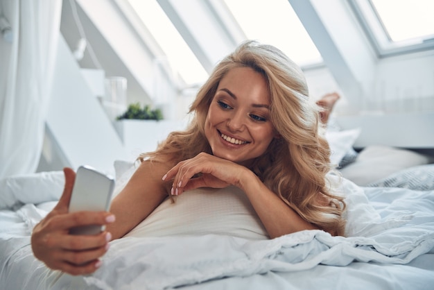 Belle jeune femme souriante utilisant un téléphone intelligent en position couchée dans son lit à la maison