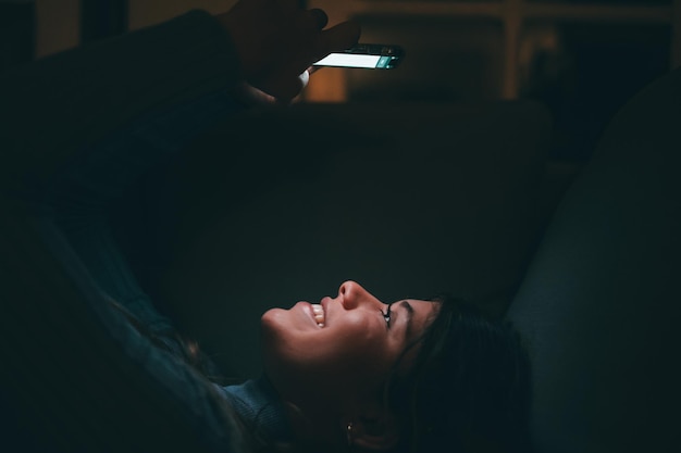 Une belle jeune femme souriante et utilisant le téléphone à la maison tard dans la nuit sur le canapé Heureuse adolescente discutant avec des amis surfant sur le net en ligne Profitant de la technologie et d'internetxA