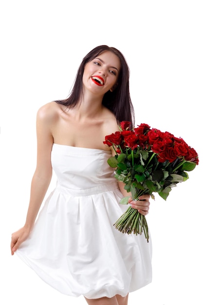 Belle jeune femme souriante vêtue d'une robe blanche avec un grand bouquet de roses rouges sur fond blanc